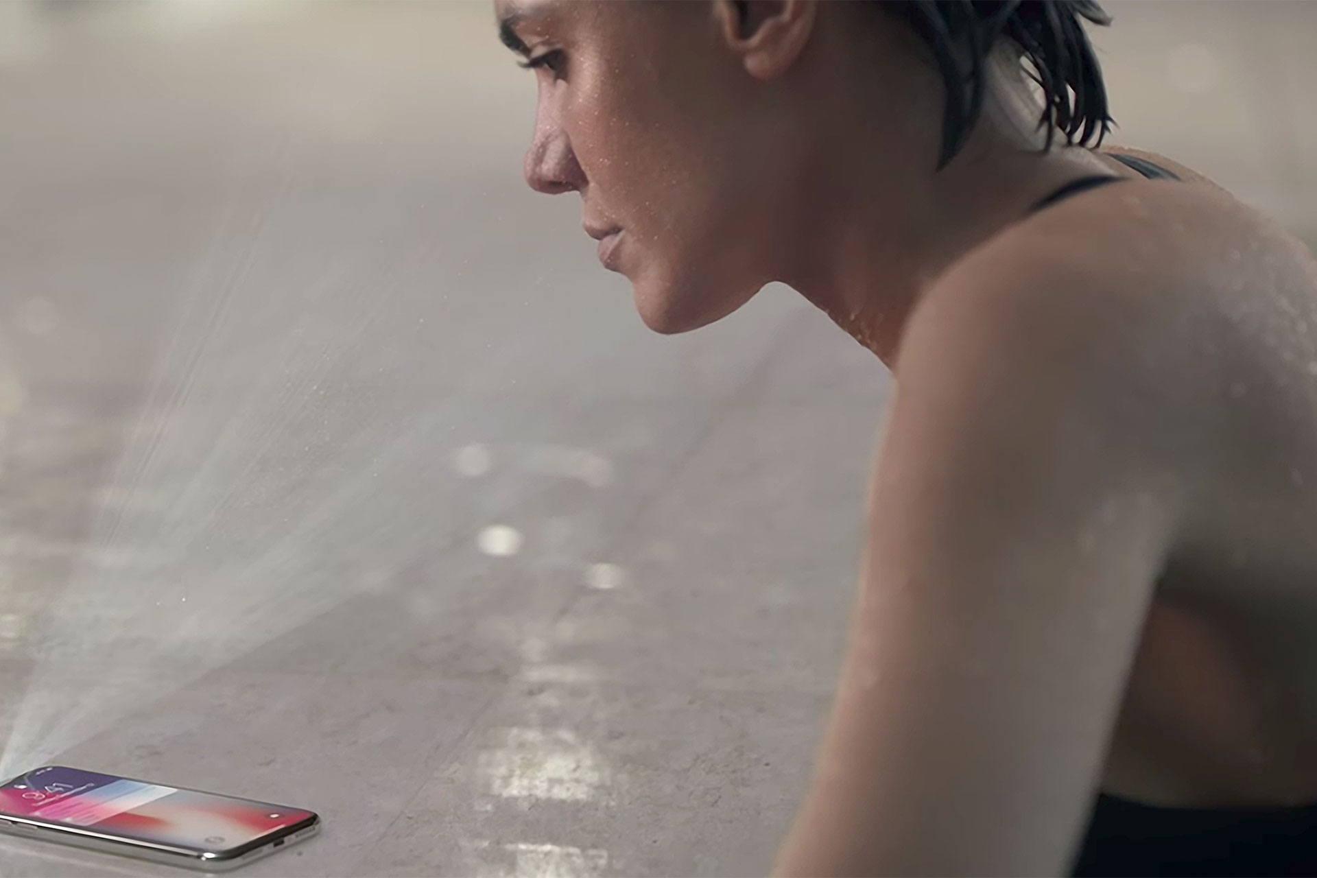A swimmer with wet hair surfacing at the edge of a pool, her face inclined toward an iPhone X resting on the wet concrete deck — a frame from Apple's 2017 Face ID advertising campaign. The phone faces her; she faces the phone.