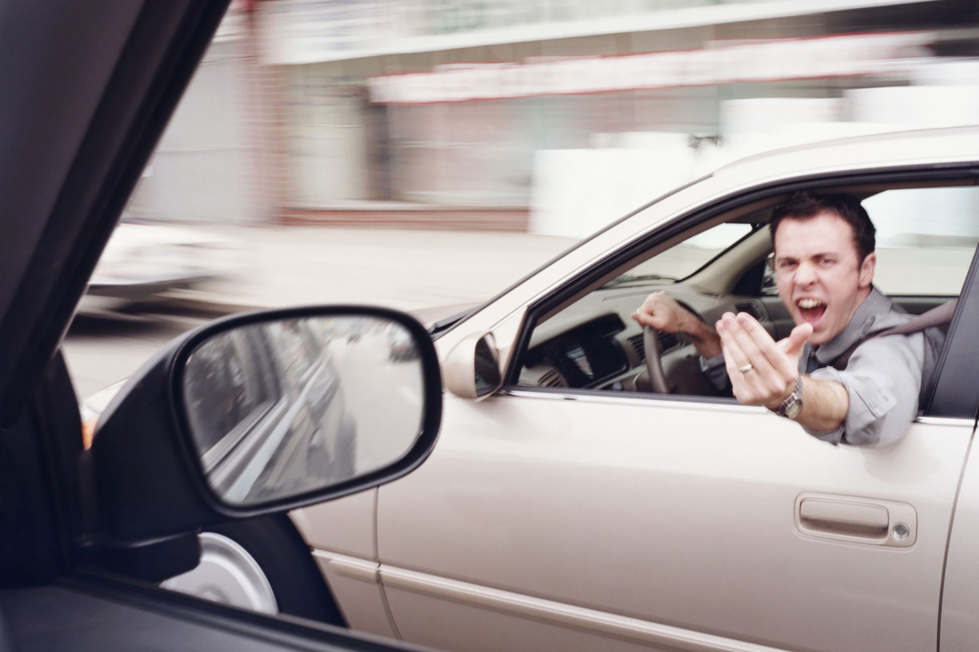 A man leans out the driver-side window of a beige sedan, mouth open in a shout, gesturing aggressively with his hand &mdash; seen through the side mirror of another car as the surrounding street blurs in motion.